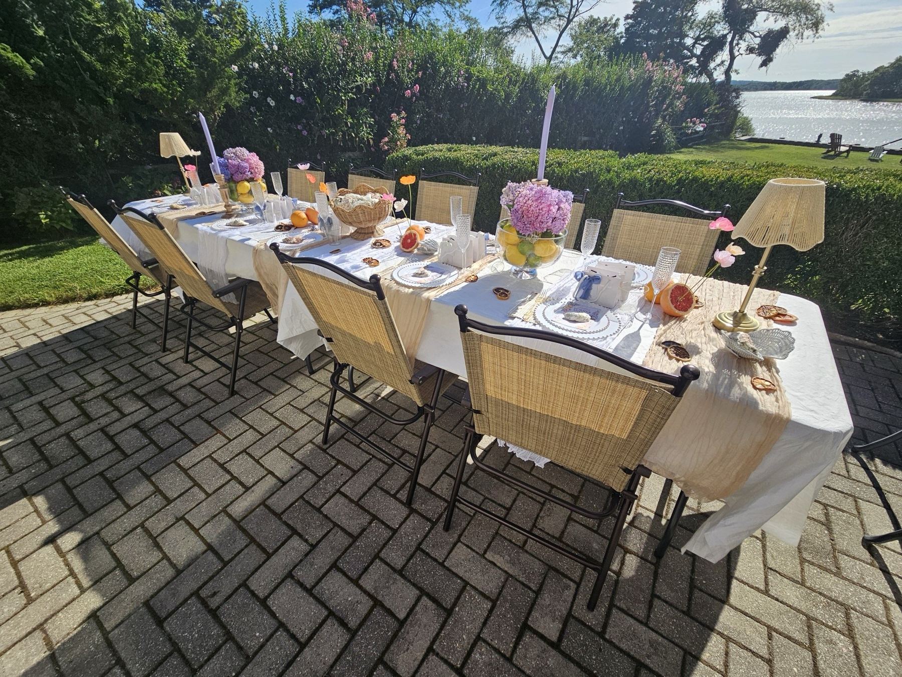The Ladies' Summer Soirée table by the water — long white table with hydrangeas, citrus, wicker lamps and lavender candles