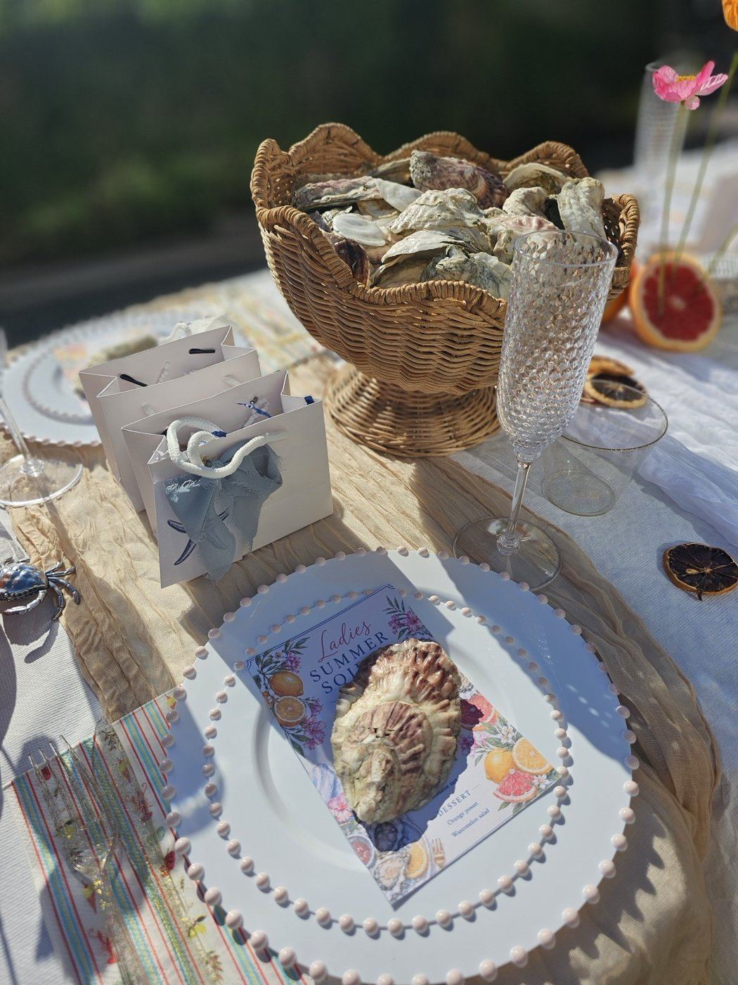 Scalloped wicker pedestal basket filled with oyster shells beside favor bags and a menu place setting